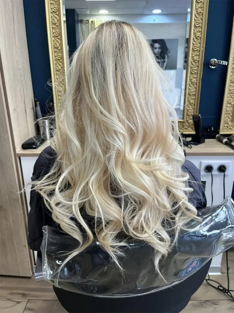 Une chevelure blonde longue et ondulée vue de dos dans un salon de coiffure.